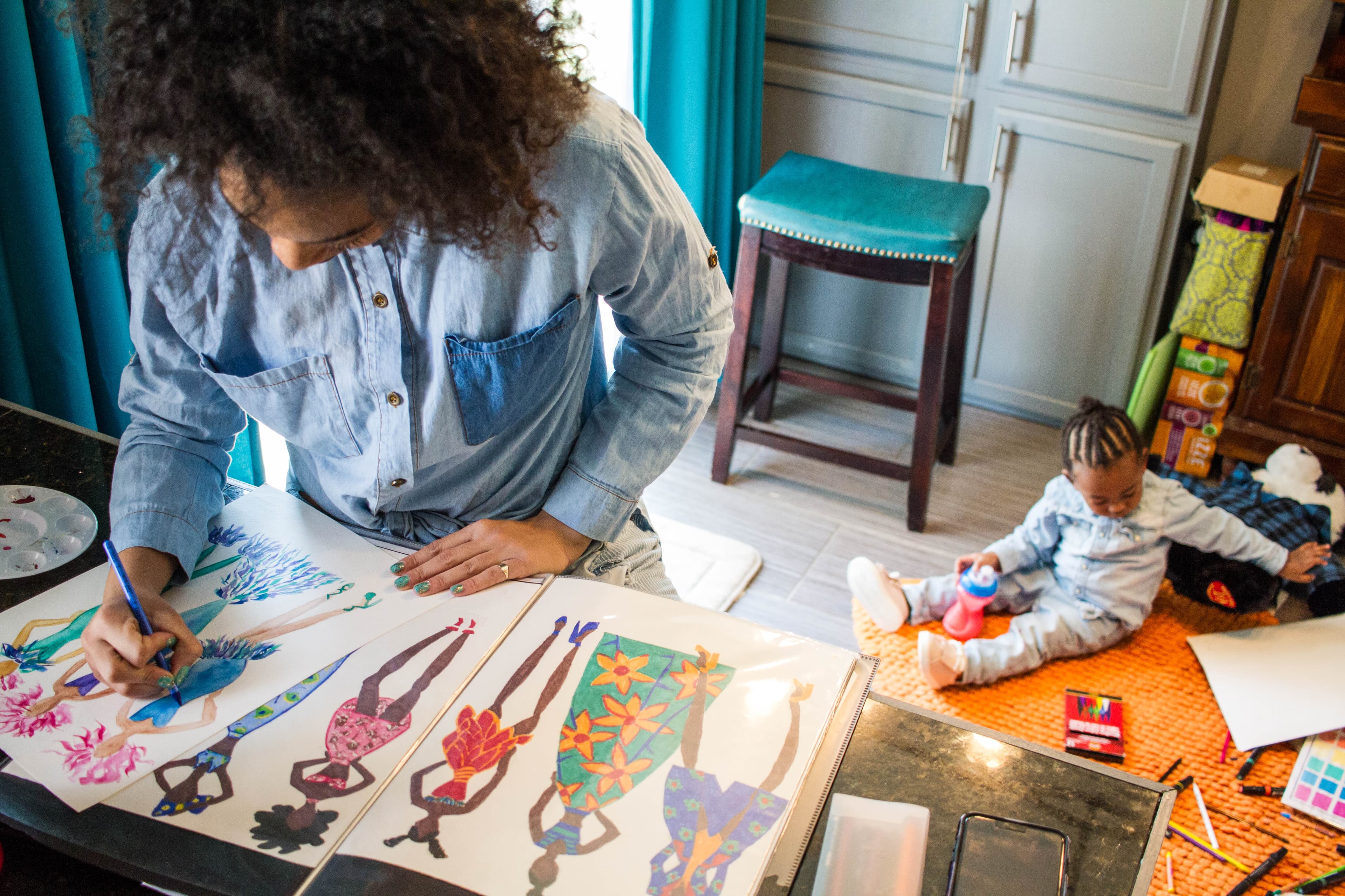 Woman standing at a desk in a bright home workspace drawing colorful fashion designs while a toddler plays with toys on the floor nearby. Woman standing at a desk in a bright home workspace drawing colorful fashion designs while a toddler plays with toys on the floor nearby.